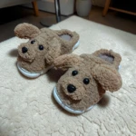 Pantuflas perrito café para niños, con suela blanca antideslizante y acolchado térmico, mostrando detalles de orejas y nariz negra sobre alfombra beige.