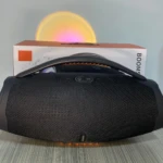 Parlante Bombox 3 negro con rejilla texturizada, luces LED en base y caja de cartón al fondo.
