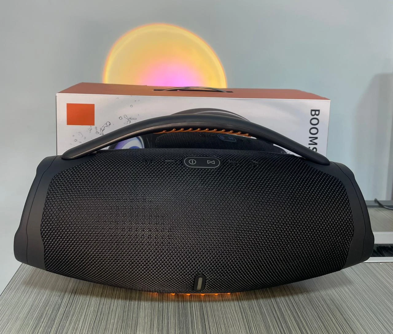Parlante Bombox 3 negro con rejilla texturizada, luces LED en base y caja de cartón al fondo.