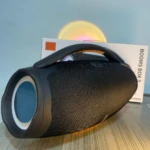Parlante portátil negro con rejilla texturizada, asa ergonómica y luces LED multicolor en la parte superior, sobre su caja original.