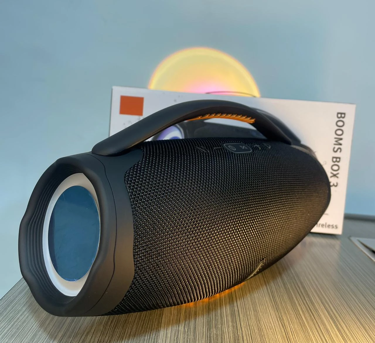 Parlante portátil negro con rejilla texturizada, asa ergonómica y luces LED multicolor en la parte superior, sobre su caja original.