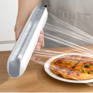 Dispensador magnético blanco con cortador invisible, extendiendo film transparente sobre pizza en plato blanco sobre mesa de madera.