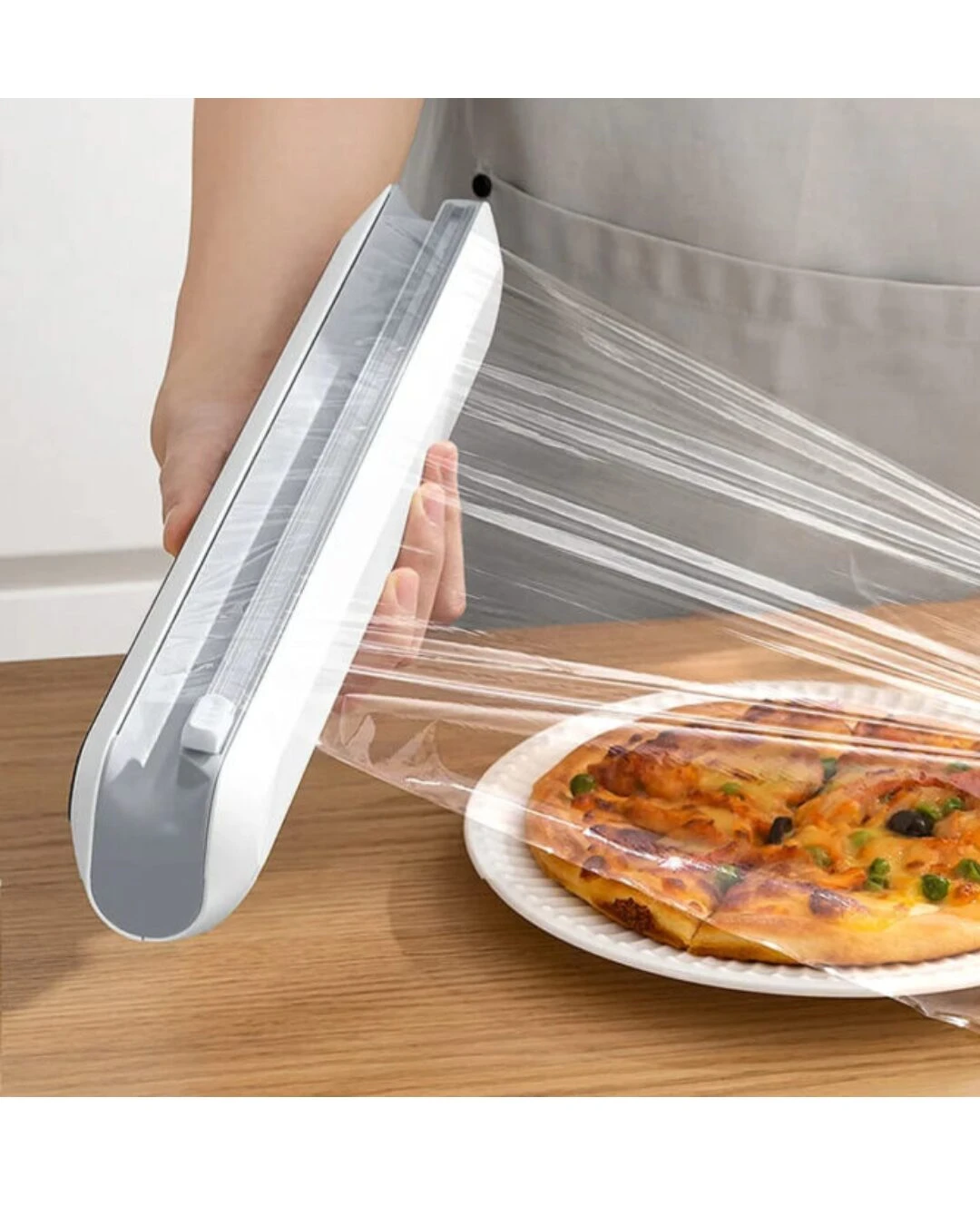Dispensador magnético blanco con cortador invisible, extendiendo film transparente sobre pizza en plato blanco sobre mesa de madera.