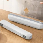 Dispensador magnético blanco con cortador invisible sobre mesa de madera, mostrando dos unidades en ángulo lateral.
