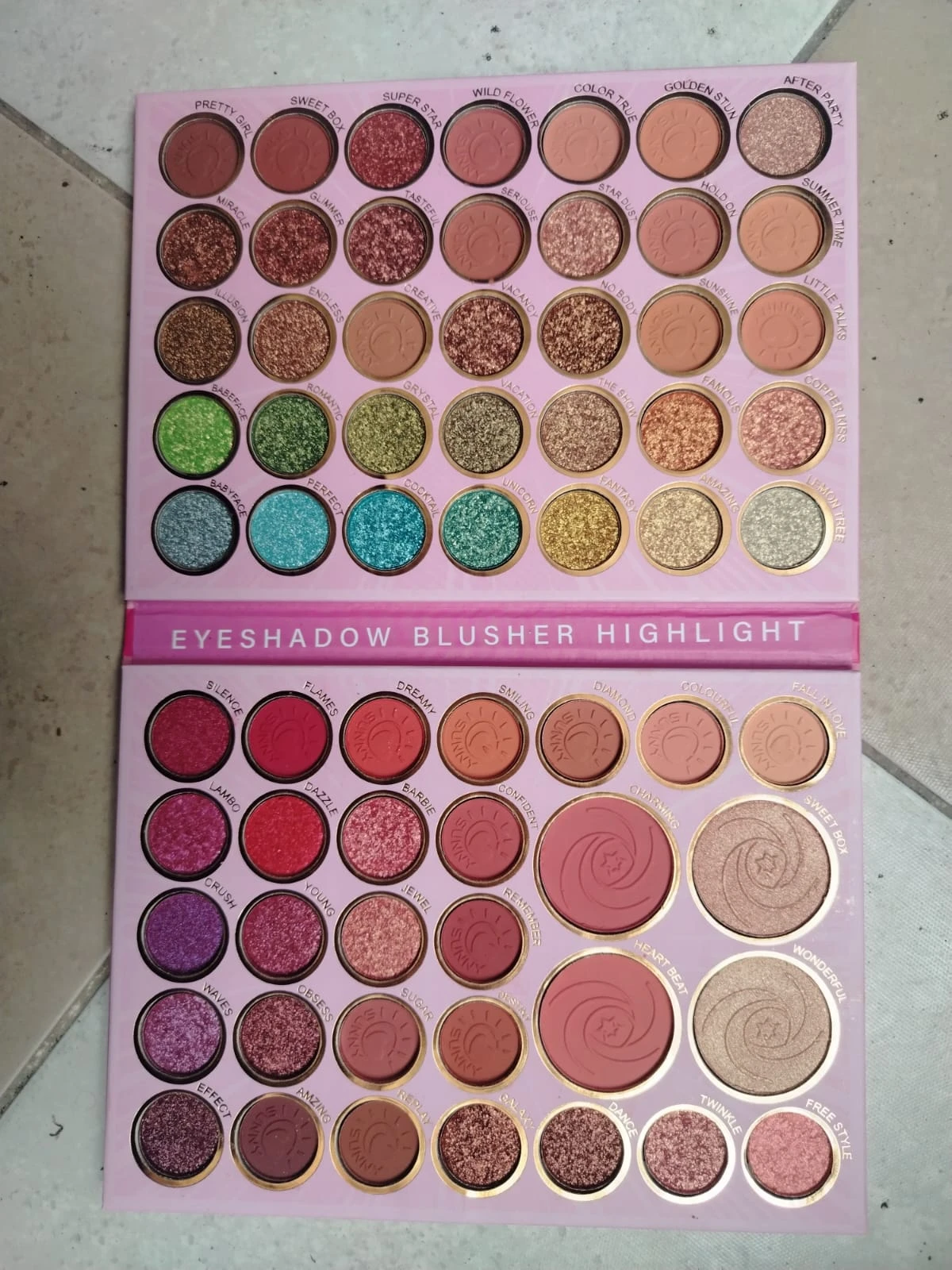 Paleta profesional de 65 sombras con tonos mate y shimmer, organizadas en tres secciones: ojos, mejillas y brillos, sobre fondo rosa claro.