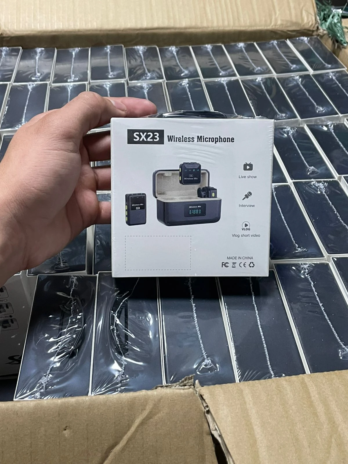 Caja blanca del micrófono inalámbrico SX23, mostrando su diseño compacto con receptor y micrófono de solapa, sobre fondo de múltiples unidades envasadas.