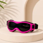 Gafas de protección para mascotas con montura rosa neón y lentes oscuras, sobre pedestal blanco con sombra de palmera al fondo.