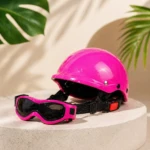Casco rosa brillante y gafas de protección con montura del mismo color, sobre pedestal circular, con hojas de planta al fondo.