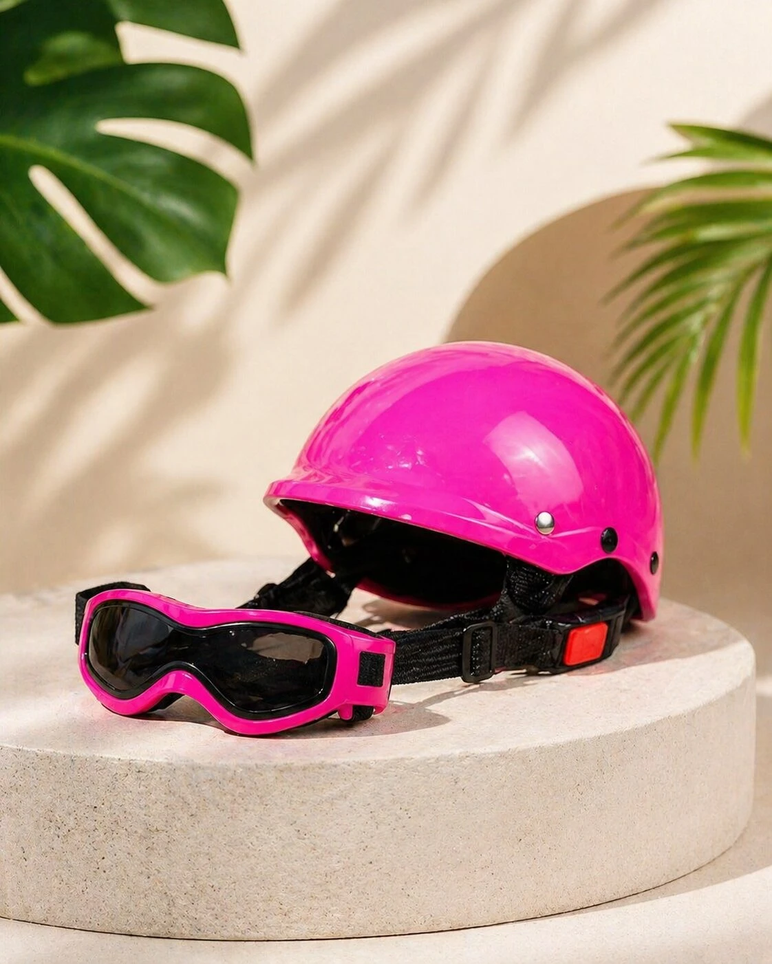 Casco rosa brillante y gafas de protección con montura del mismo color, sobre pedestal circular, con hojas de planta al fondo.