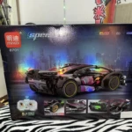 Caja del set de construcción Mingdi 8701, mostrando un coche deportivo negro con detalles multicolores y luces LED, sobre fondo urbano nocturno. Incluye mando a distancia y accesorios visibles en la parte inferior.