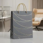 Bolsa regalo mediana mate gris con líneas doradas y asas trenzadas, sobre superficie blanca en interior moderno.