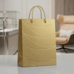 Bolsa de regalo mediana color mate dorado con textura ondulada y asas trenzadas, sobre superficie blanca en interior moderno.