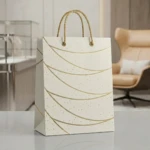 Bolsa regalo mediana color crema con detalles dorados y asas trenzadas, sobre superficie blanca en interior moderno.