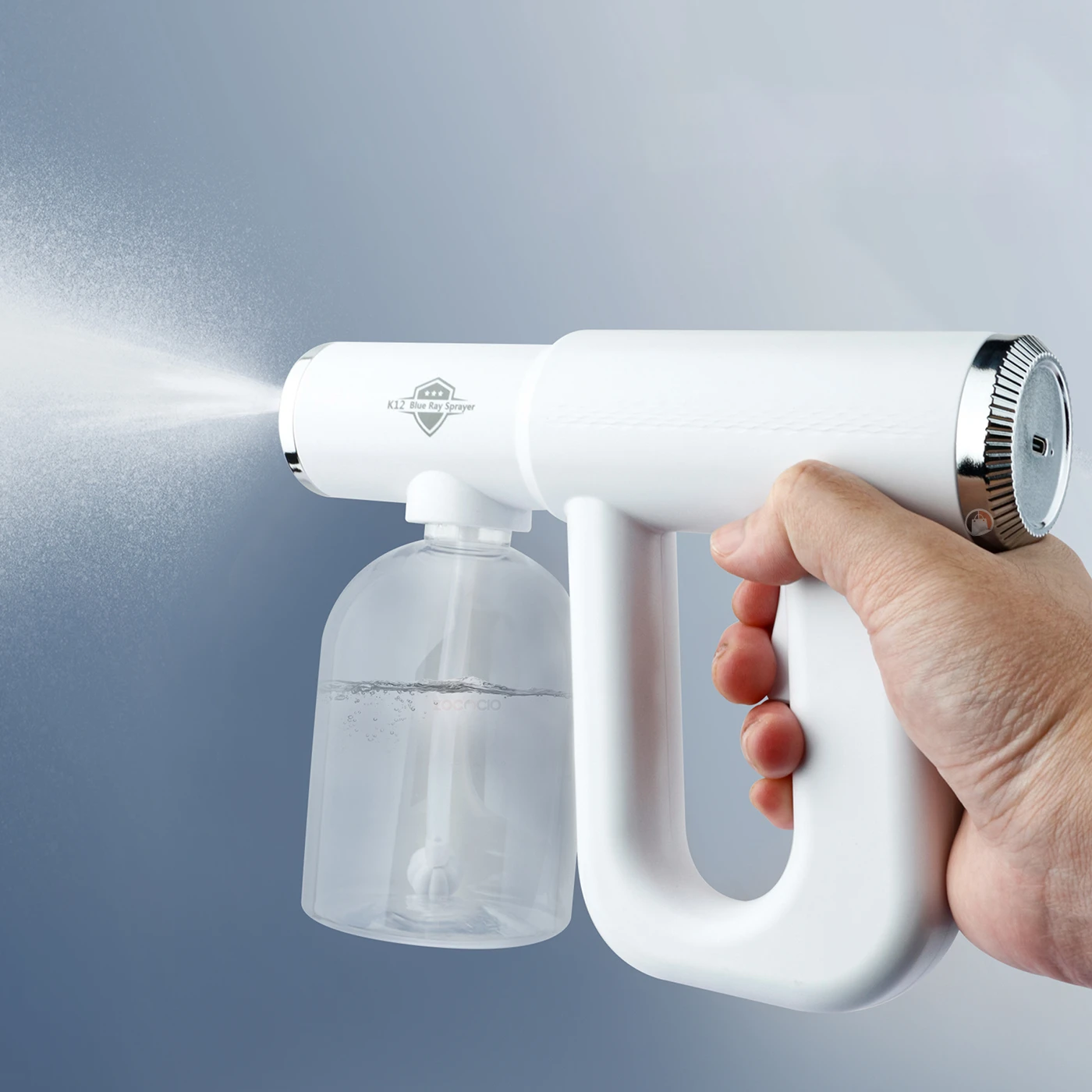 Pistola pulverizadora blanca con depósito transparente, accionada por mano, emitiendo niebla fina sobre superficie gris.