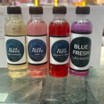 Cuatro botellas de esencia Blue Fresh de 60ml en colores amarillo, rosa, rojo y morado, con etiquetas que indican sabores como coco vainilla, yara, flor de hibisco y lavanda.