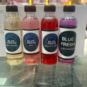 Cuatro botellas de esencia Blue Fresh de 60ml en colores amarillo, rosa, rojo y morado, con etiquetas que indican sabores como coco vainilla, yara, flor de hibisco y lavanda.