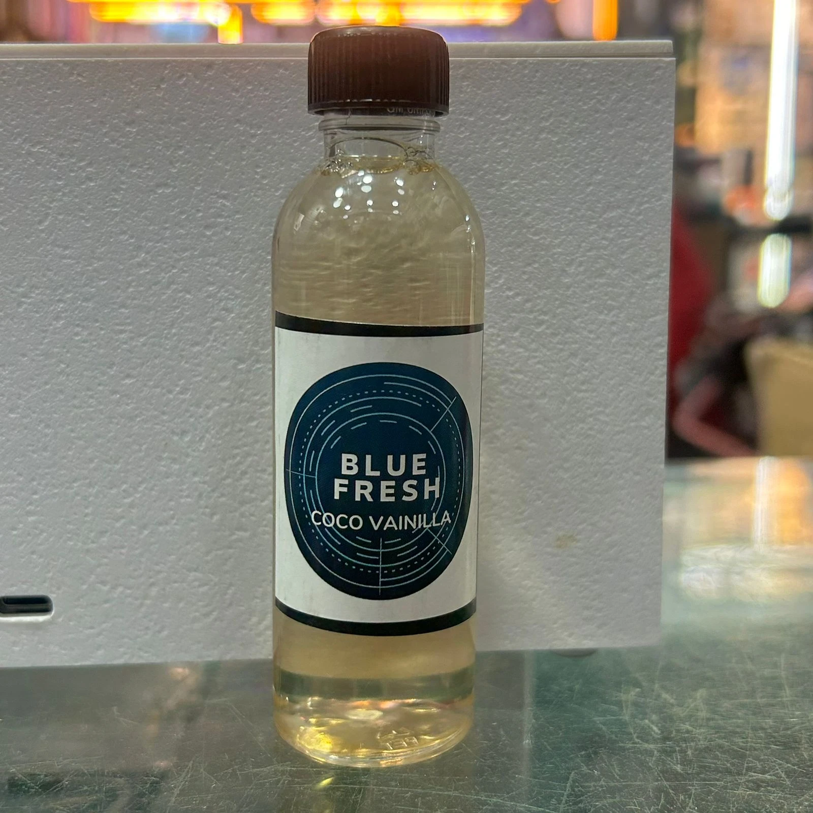 Bote de 60 ml de esencia Blue Fresh sabor coco vainilla, con etiqueta circular azul y tapa marrón, sobre superficie metálica.