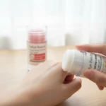 Crema hidratante para manos y pies en presentación de barra, con textura blanca y aplicador de silicona, siendo usada sobre la piel de una mano. En segundo plano, otro envase del mismo producto con etiqueta visible.