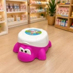 Orinal en forma de tortuga fucsia con tapa blanca y diseño de Doraemon, sobre piso de madera en tienda de artículos infantiles.