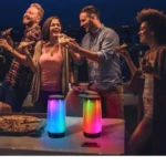 Dos parlantes Remax con luces LED multicolor en mesa, rodeados de amigos brindando con pizza al aire libre.