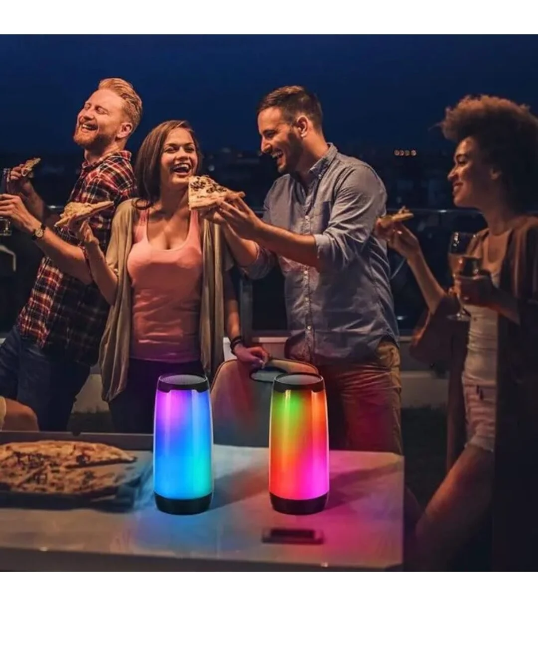 Dos parlantes Remax con luces LED multicolor en mesa, rodeados de amigos brindando con pizza al aire libre.