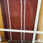 Armario de lona marrón con cinco compartimentos y zona de colgar, sobre ruedas, en ambiente interior.