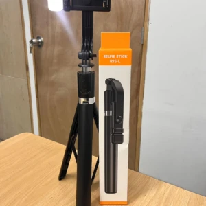 Tripode selfie negro con luz LED frontal y control remoto, desplegado sobre mesa de madera junto a su caja naranja.