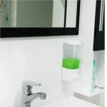 Dispensador de jabón líquido transparente con cuerpo blanco, contenido verde, montado en pared junto a lavabo y espejo.