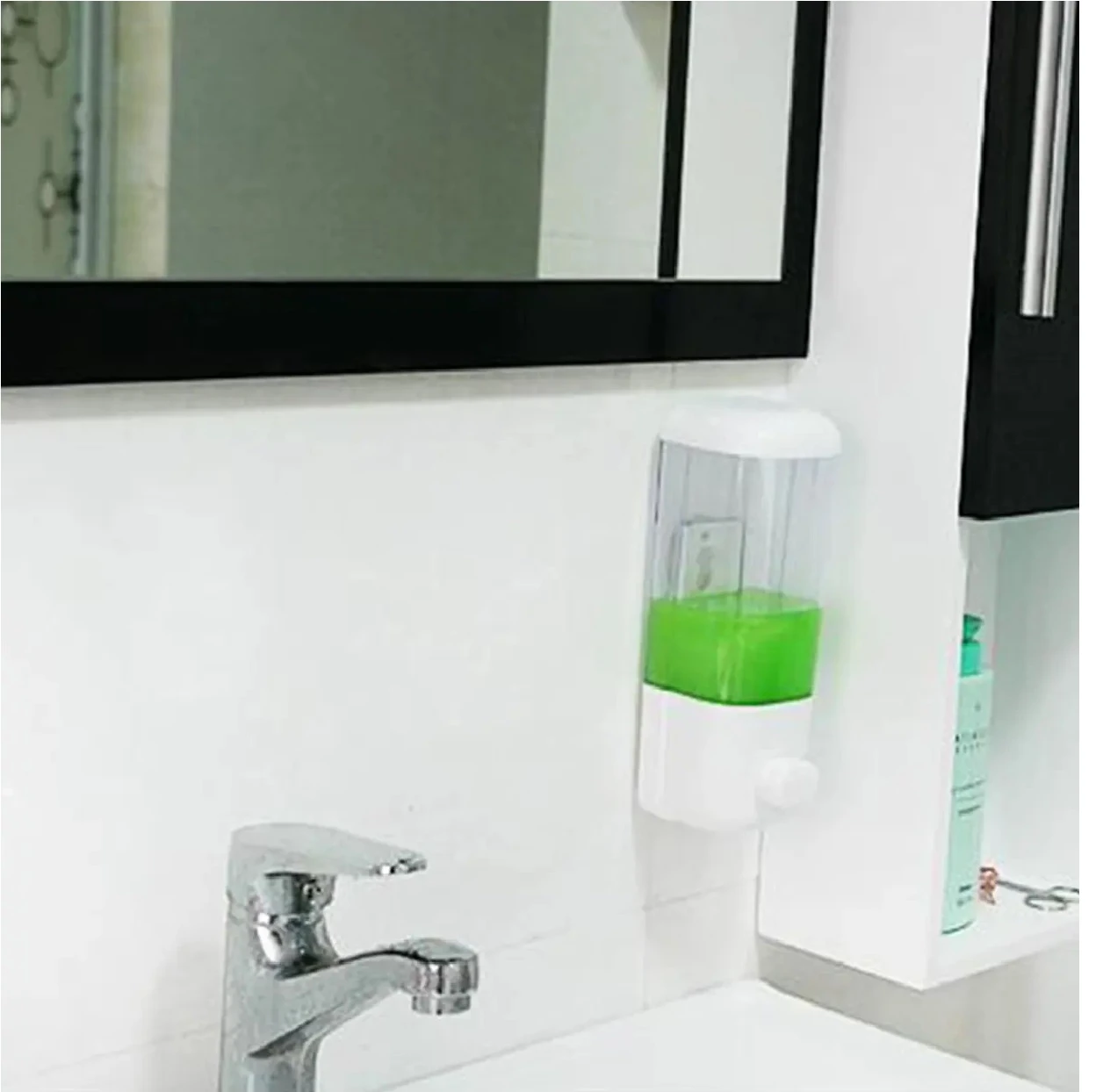 Dispensador de jabón líquido transparente con cuerpo blanco, contenido verde, montado en pared junto a lavabo y espejo.