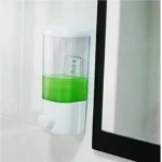 Dispensador de jabón líquido transparente con cuerpo blanco, contenido verde visible, montado en pared junto a espejo negro.