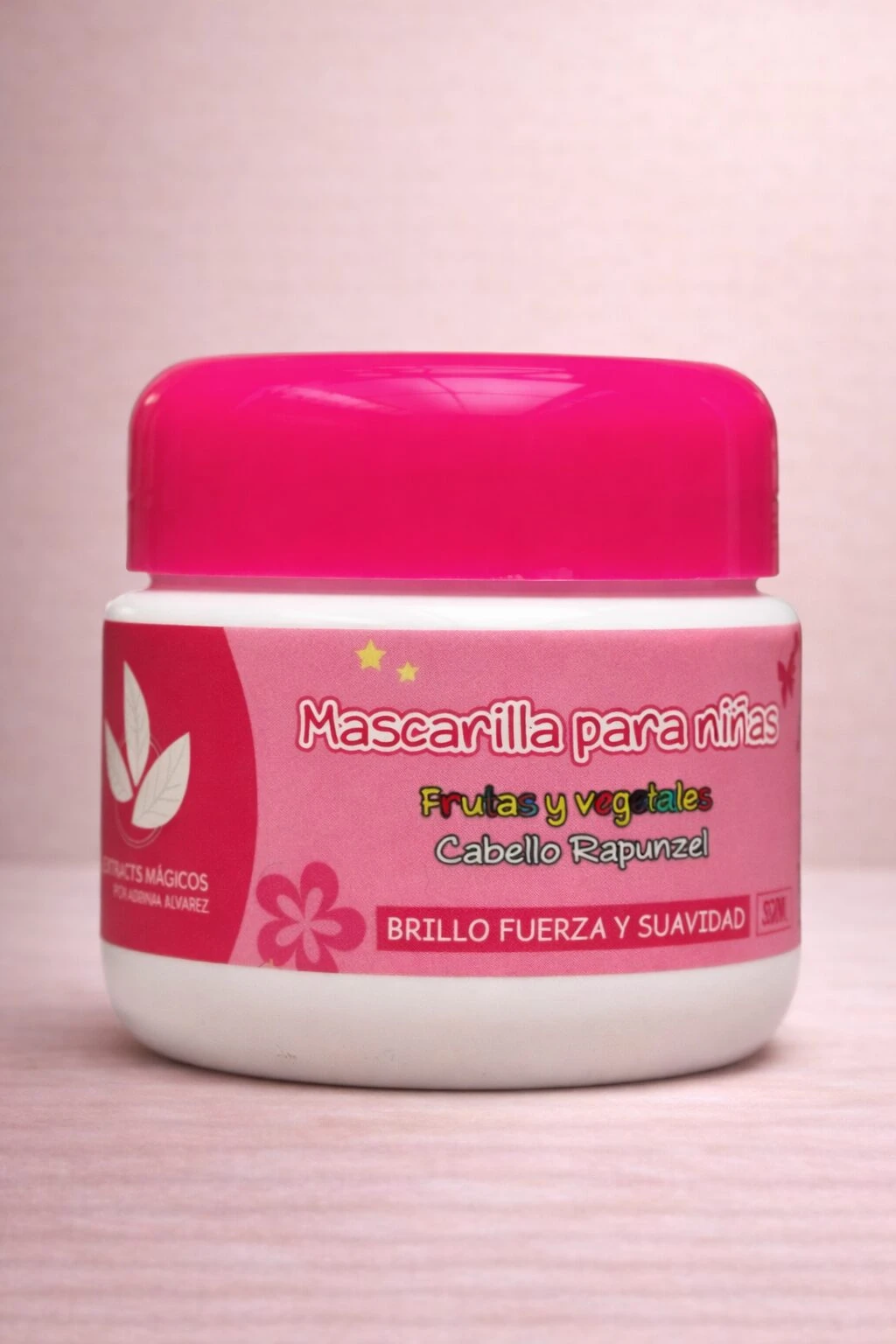Jarrón blanco con tapa rosa brillante, etiqueta con diseño floral y texto en español que indica mascarilla capilar para niñas, con frutas y vegetales, cabello Rapunzel, brillo, fuerza y suavidad.