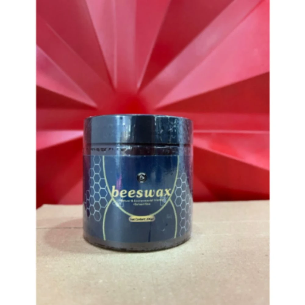 Cera de miel protectora de muebles en frasco azul oscuro con tapa negra, etiqueta con diseño de panal y texto visible. Producto sobre superficie clara con fondo rojo geométrico.