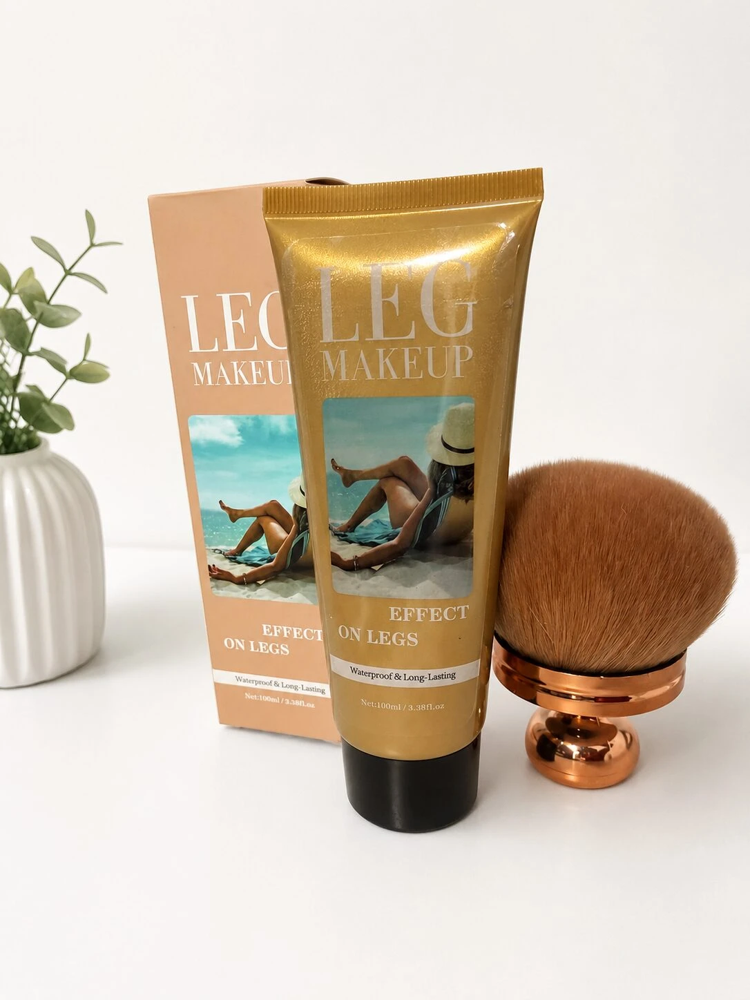 Tubo dorado de maquillaje para piernas con aplicador resistente al agua, acompañado de cepillo y caja, sobre fondo blanco con planta decorativa.