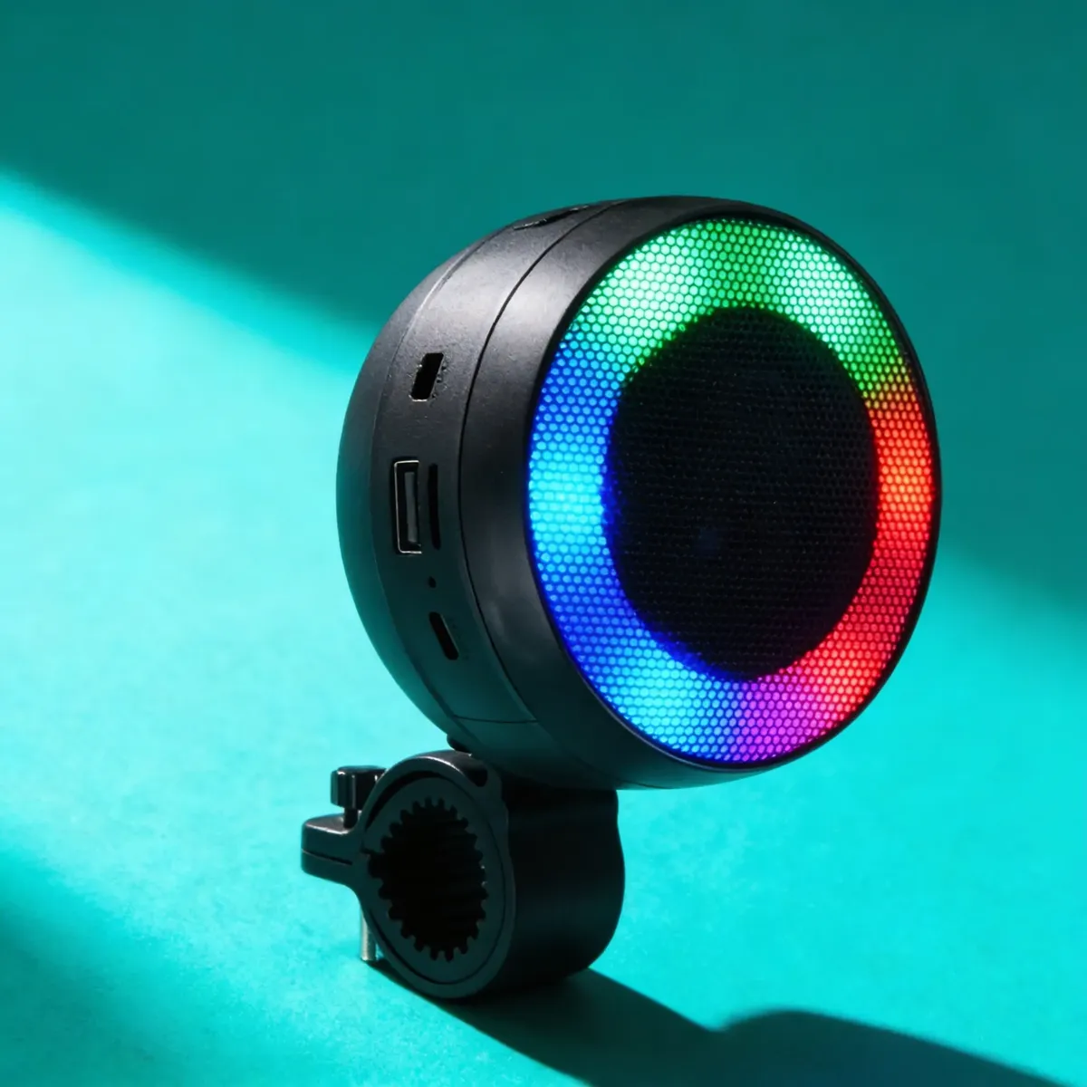Parlante Bluetooth negro con luces LED RGB en anillo, montado en soporte para manillar de bicicleta, sobre fondo turquesa.