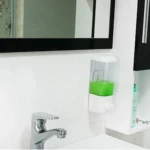 Dispensador de jabón líquido transparente de 500 ml en ABS, con líquido verde, montado en pared junto a lavabo y espejo.