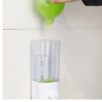 Dispensador de jabón líquido transparente de 500 ml en ABS, con tapa blanca y dispensador en uso, viéndose jabón verde siendo vertido desde un envase.