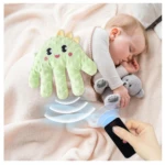 Almohada Mano Arrulla en color verde menta, con diseño de mano y rostro sonriente, emitiendo ondas de sonido visibles. Bebé durmiendo junto a peluche gris, control remoto negro en mano adulta sobre manta rosa.