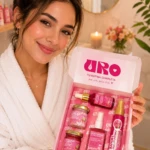 Mujer sonriendo en bata blanca sosteniendo caja rosa abierta con productos de cuidado íntimo y colágeno, incluyendo frascos, tubos y botellas con etiquetas visibles.