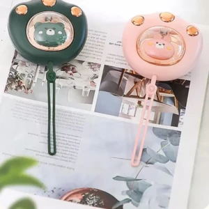 Dos mini calentadores de manos en forma de garra, uno verde y otro rosa, con diseño de oso y detalles dorados, sobre revista abierta.