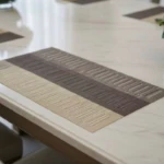 Tapete rectangular de mesa bicolor en tonos gris y beige, con textura de líneas horizontales, colocado sobre superficie blanca brillante en ambiente de comedor moderno.