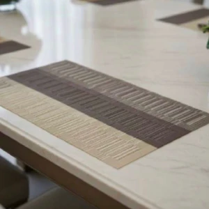 Tapete rectangular de mesa bicolor en tonos gris y beige, con textura de líneas horizontales, colocado sobre superficie blanca brillante en ambiente de comedor moderno.