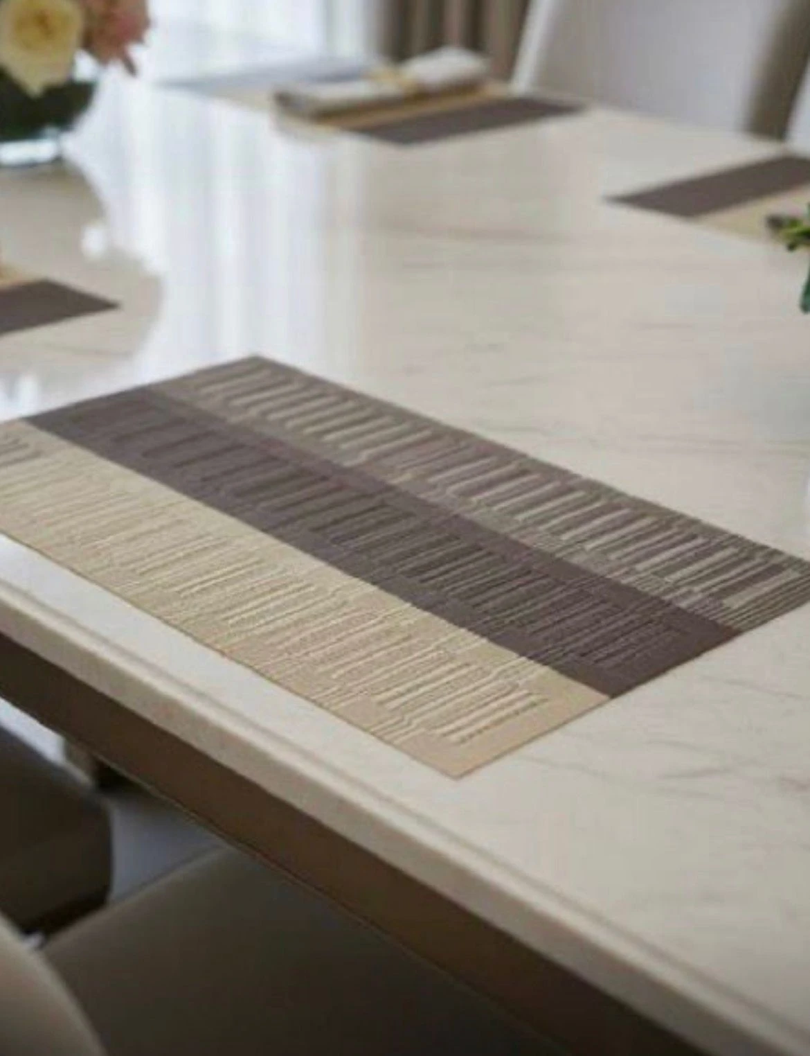 Tapete rectangular de mesa bicolor en tonos gris y beige, con textura de líneas horizontales, colocado sobre superficie blanca brillante en ambiente de comedor moderno.