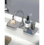 Almohadilla de drenaje en silicona gris bajo grifo, con soporte para jabonera, cepillo y vaso, sobre encimera blanca.