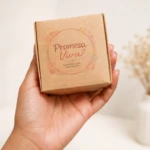 Caja de cartón marrón con diseño circular en tonos cálidos que dice Promesa Viva y Tarjetas con Propósito, sostenida en mano sobre fondo blanco con ramos secos al fondo.