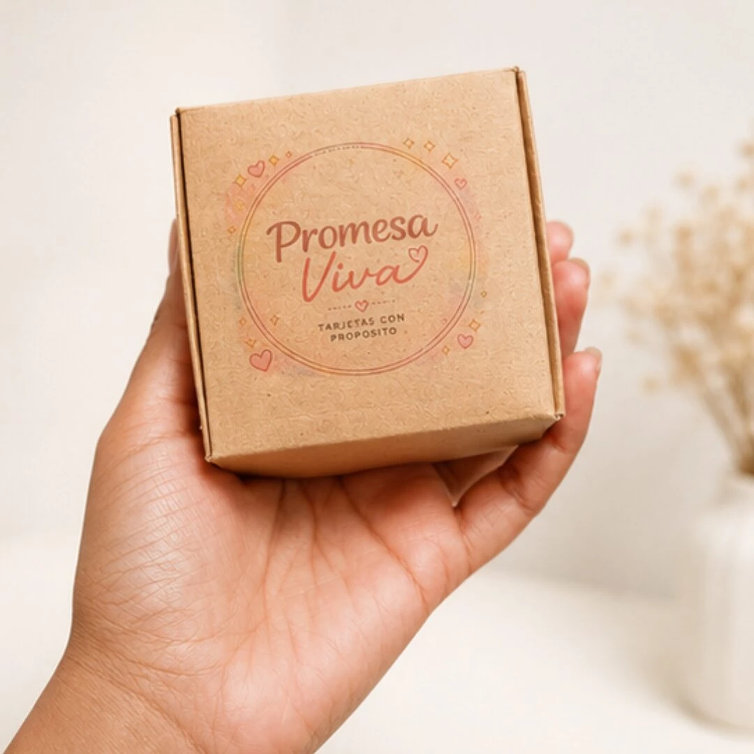 Caja de cartón marrón con diseño circular en tonos cálidos que dice Promesa Viva y Tarjetas con Propósito, sostenida en mano sobre fondo blanco con ramos secos al fondo.