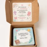 Caja de cartón abierta mostrando tarjetas con mensajes bíblicos, diseño pastel con ilustraciones de arcoíris, cruz y libro, texto visible en tapa y tarjeta interior.