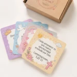Tarjetas con mensajes bíblicos Promesa V, diseño cuadrado en colores pastel con ilustraciones de sol, cruz, paloma y arcoíris, junto a caja de cartón marrón.