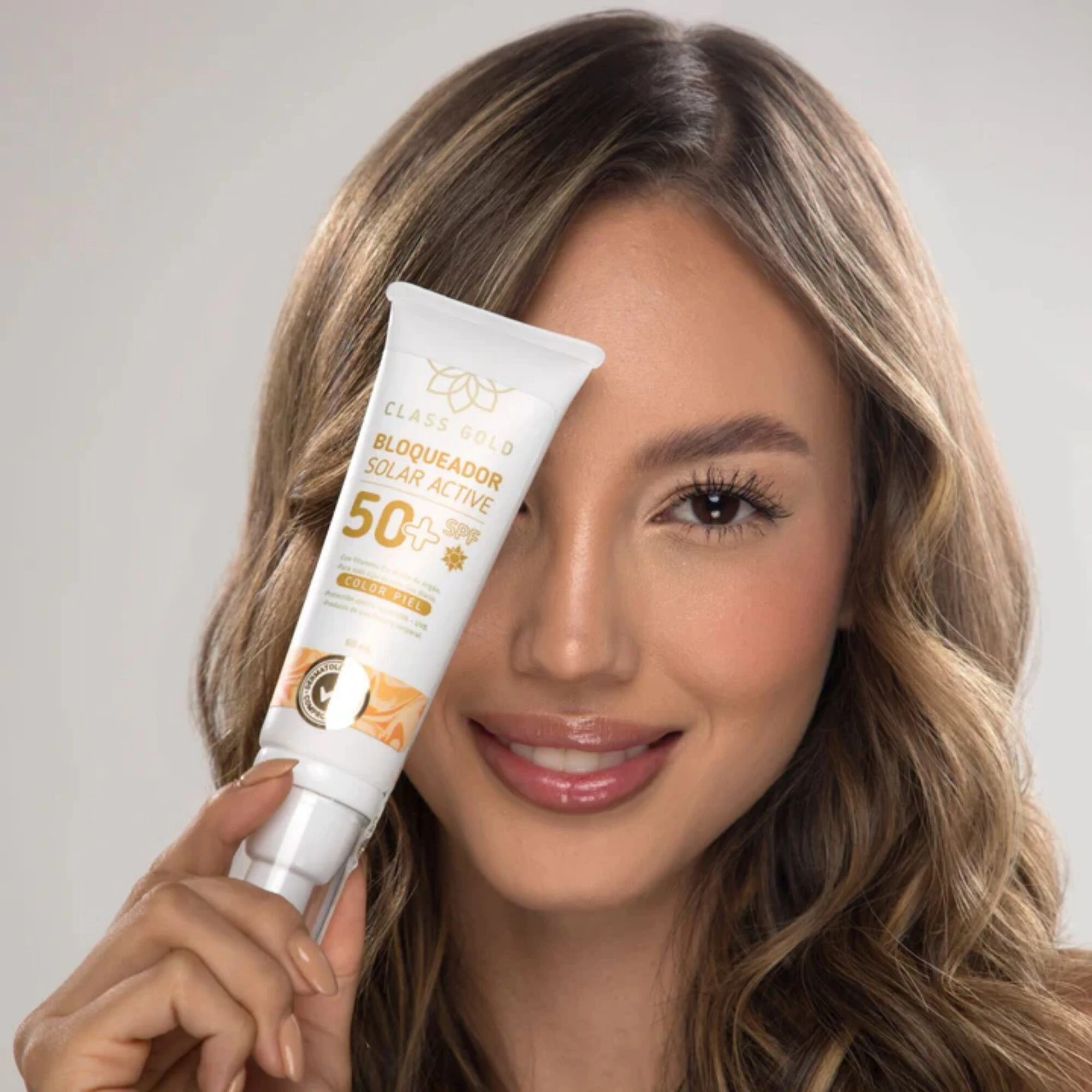 Mujer sonriendo mostrando tubo blanco de bloqueador solar facial SPF 50 con pigmentos, aplicando producto en rostro para uniformizar tono.