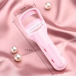 Rizador de pestañas manual en color rosa pastel, con mango ergonómico y medidas de 11 cm de largo y 4 cm de ancho, sobre fondo de tela rosa con perlas decorativas.