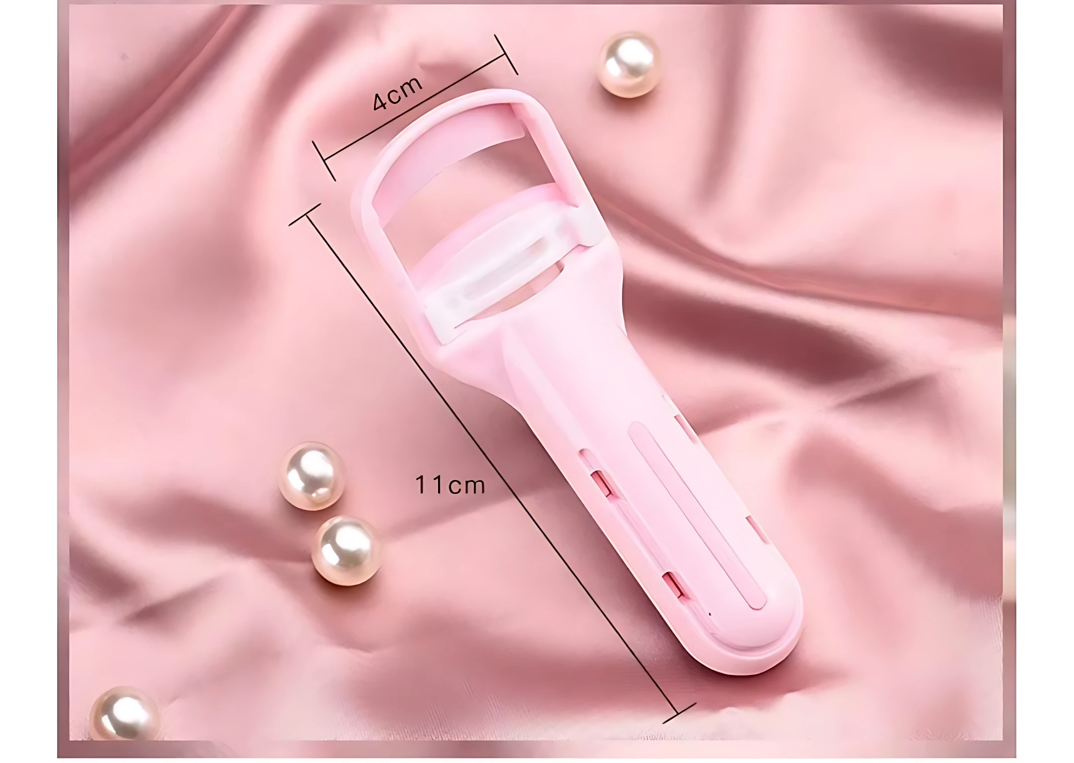 Rizador de pestañas manual ergonómico en color rosa pastel, con medidas de 11 cm de largo y 4 cm de ancho, sobre tela rosa con perlas decorativas.